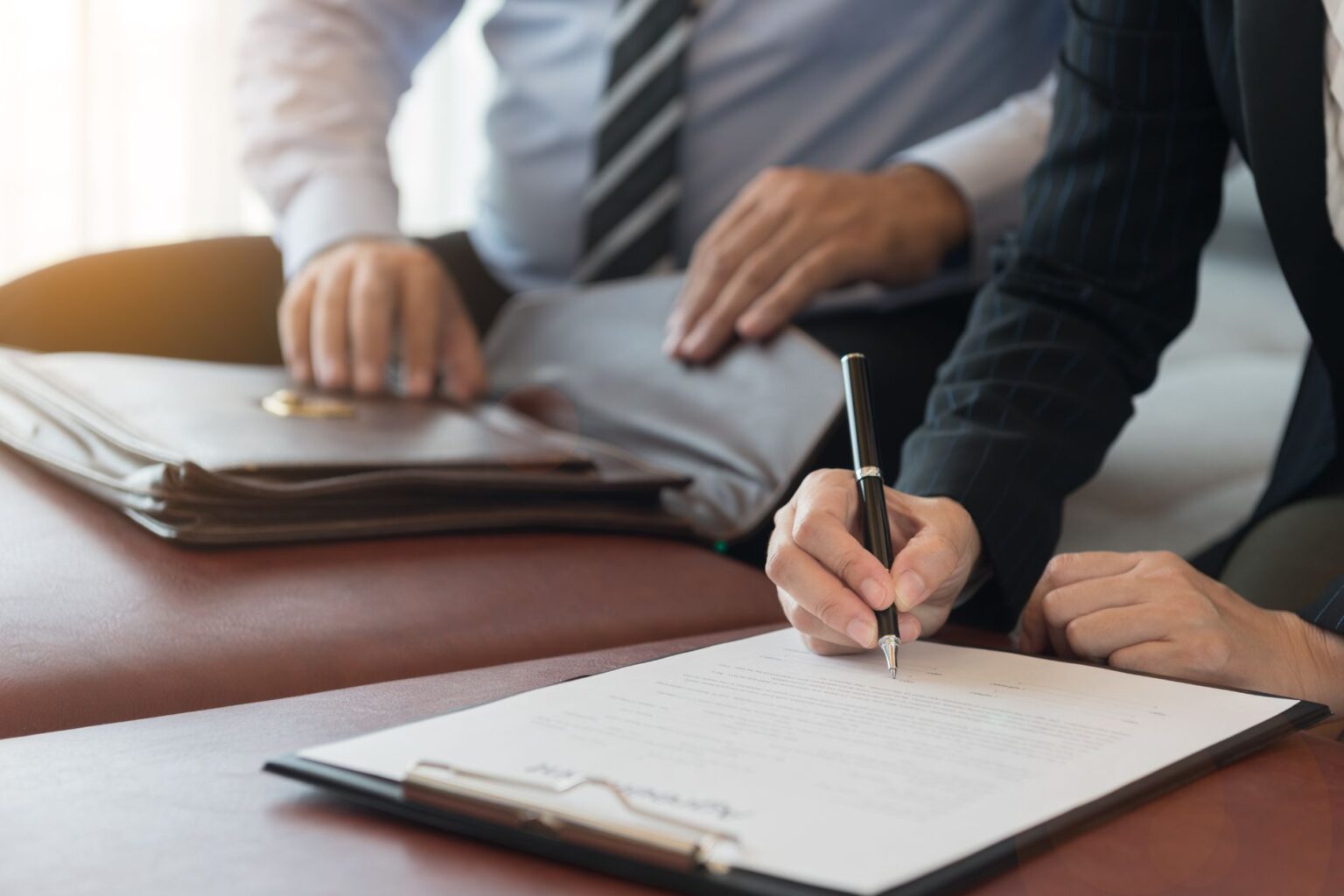 a man wearing a suit signs an agreement document, in front of another suited man holding a briefcase