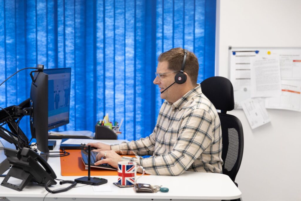 Jon is pictured smiling in business casual wear, at his office desk with a headset on